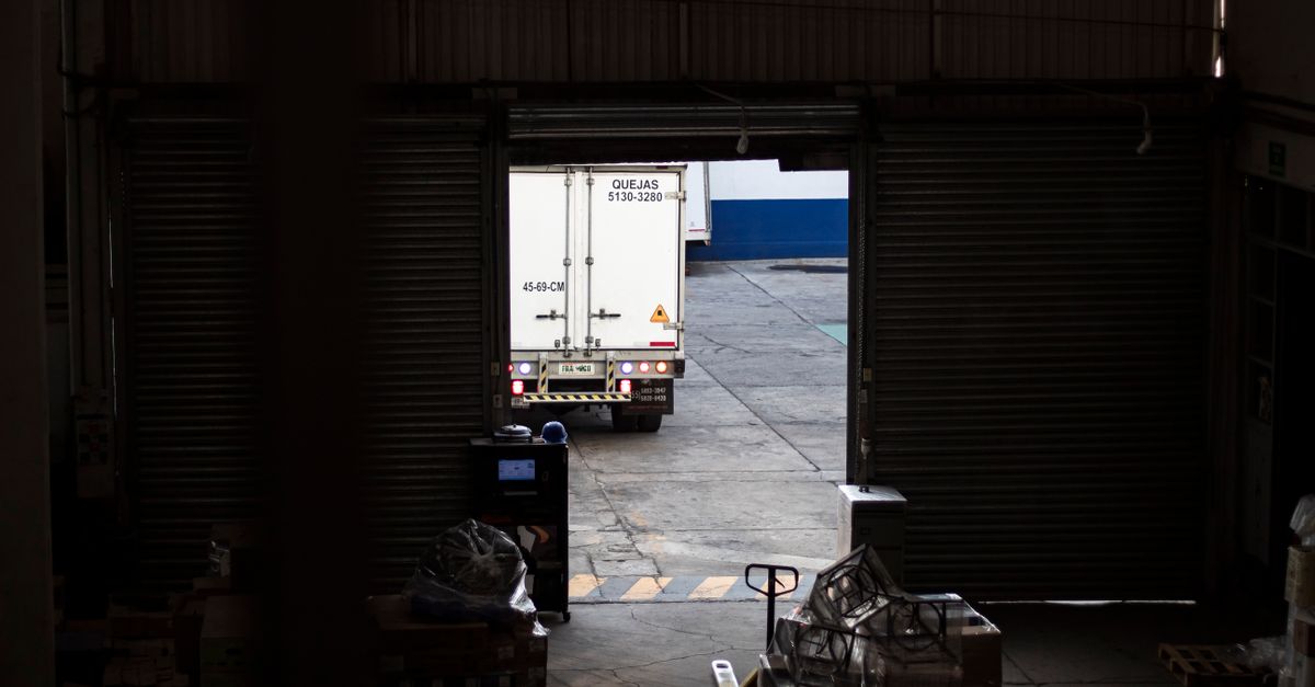A white delivery truck exits a dimly lit warehouse loading dock, surrounded by packages and pallets.